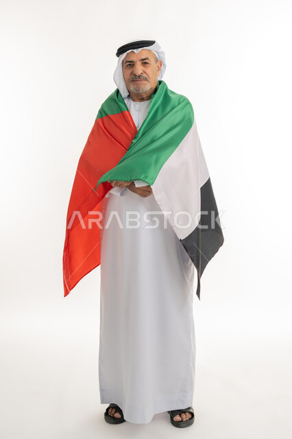 النظر الى الكاميرا بايماءات الاعتزاز والثقة، هوية اليوم الوطني الاماراتي 51، احياء ذكرى استقلال الإمارات، بورتريه لرجل مسن عربي خليجي إماراتي يرتدي الكندورة والغترة يقوم بحمل علم الإمارات على ظهره، الحب والانتماء للوطن، الطول الكامل للجسم، خلفية بيضاء
