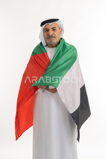 احياء ذكرى استقلال الإمارات، بورتريه لرجل مسن عربي خليجي إماراتي يرتدي الكندورة والغترة يقوم بحمل علم الإمارات العربية المتحدة على ظهره، الحب والانتماء للوطن، النظر الى الكاميرا بايماءات الاعتزاز والثقة، هوية اليوم الوطني الاماراتي 51، خلفية بيضاء