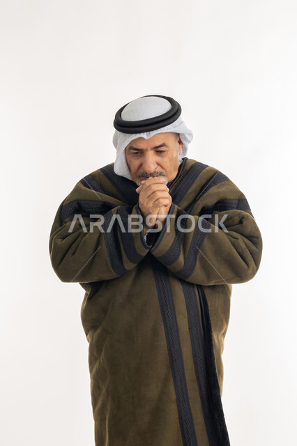 الشعور بالصقيع والقشعريرة والتجمد، إيماءات وجه ويدين تدل على البرد الشديد، بورتريه لرجل مسن عربي خليجي اماراتي  يرتدي الفروة والغترة التقليدية يقوم بتدفئة يديه، ارتداء الملابس الدافئة لكبار السن، الأجواء الشتوية الباردة، خلفية بيضاء