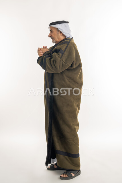 إيماءات وجه ويدين تدل على البرد الشديد، بورتريه من الجانب لرجل مسن عربي خليجي اماراتي  يرتدي الفروة والغترة التقليدية يقوم بتدفئة يديه، ارتداء الملابس الدافئة لكبار السن، الأجواء الشتوية الباردة، الشعور بالصقيع والقشعريرة والتجمد، الطول الكامل للجسم، خلفية بيضاء