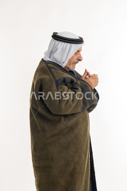 ارتداء الملابس الدافئة لكبار السن، الأجواء الشتوية الباردة، الشعور بالصقيع والقشعريرة والتجمد، إيماءات وجه ويدين تدل على البرد الشديد، بورتريه من الجانب لرجل مسن عربي خليجي اماراتي  يرتدي الفروة والغترة التقليدية يقوم بتدفئة يديه، خلفية بيضاء