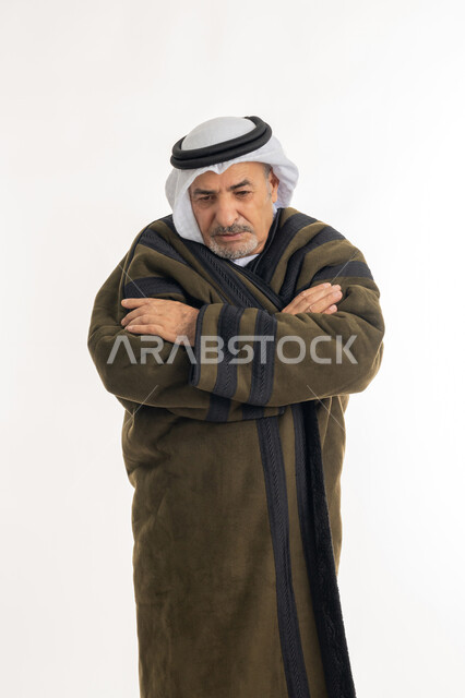 Freezing and feeling frosty and chills, Portrait of an elderly Arab Gulf Emirati man wearing a fur and ghutra standing with his hands folded, Importance of wearing warm clothes for the elderly in winter, Facial and hand gestures indicating extreme cold, White background
