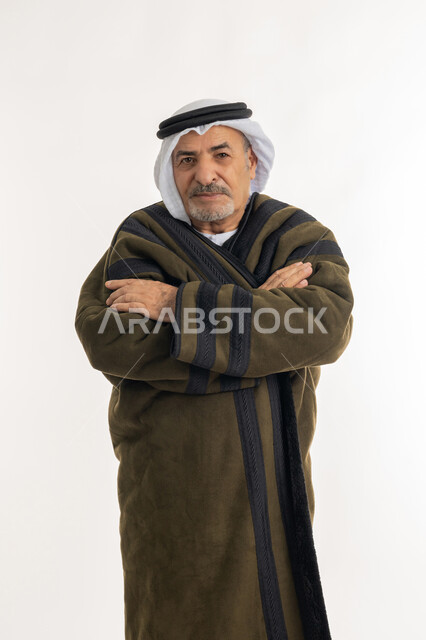 النظر الى الكاميرا بايماءات السرور، البرودة الشديدة لفصل الشتاء، بورتريه لرجل مسن عربي خليجي اماراتي  يرتدي الفروة والغترة يقف مكتوف الأيدي، الشعور بالصقيع والقشعريرة والتجمد، إيماءات وجه ويدين تدل على البرد الشديد، خلفية بيضاء