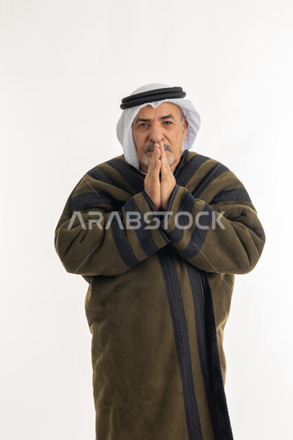 أهمية ارتداء الملابس الدافئة لكبار السن في الأجواء الشتوية، بورتريه لرجل مسن عربي خليجي اماراتي  يرتدي الفروة والغترة التقليدية يقوم بتدفئة يديه، النظر إلى الكاميرا بإيماءات السرور والثقة بالنفس، الشعور بالبرد القارس في فصل الشتاء، خلفية بيضاء