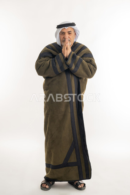 الشعور بالصقيع والقشعريرة والتجمد، أهمية ارتداء الملابس الدافئة لكبار السن في الأجواء الشتوية، بورتريه لرجل مسن عربي خليجي اماراتي  يرتدي الفروة والغترة يقوم بتدفئة يديه، النظر إلى الكاميرا بإيماءات السرور والثقة بالنفس، الطول الكامل للجسم، خلفية بيضاء