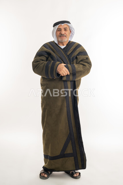 النظر إلى الكاميرا بإيماءات السرور والثقة بالنفس، الشعور بالبرد القارس في فصل الشتاء، أهمية ارتداء الملابس الدافئة لكبار السن في الأجواء الشتوية، بورتريه لرجل مسن عربي خليجي اماراتي  يرتدي الفروة والغترة التقليدية، صورة الجسم بالكامل، خلفية بيضاء