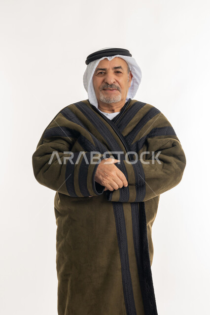 الشعور بالبرد القارس في فصل الشتاء، النظر إلى الكاميرا بإيماءات السرور والثقة بالنفس، أهمية ارتداء الملابس الدافئة لكبار السن في الأجواء الشتوية، بورتريه لرجل مسن عربي خليجي اماراتي  يرتدي الفروة والغترة التقليدية، خلفية بيضاء