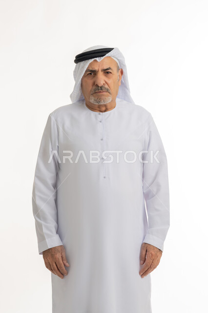 النظر الى الكاميرا بايماءات السخط والتذمر، مفهوم عدم القبول والرضا، الشعور بالعصبية والضجر، تعابير العبوس والانزعاج، نظرات التحدي والغضب، بورتريه لرجل عربي خليجي إماراتي كبير بالسن يرتدي الغترة والكندورة الإماراتية، خلفية بيضاء