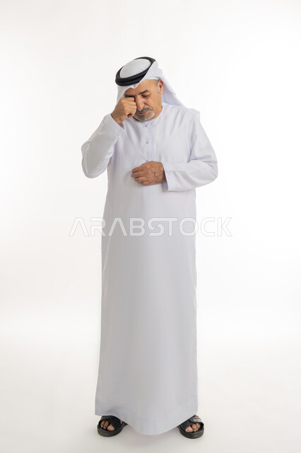 الوقوف والشعور بالاضطراب والتعب، إشارات وحركات وجه ويدين مختلفة تدل على الانزعاج وعدم الرضا، بورتريه لرجل عربي خليجي إماراتي كبير بالسن يرتدي الكندورة الإماراتية التقليدية والغترة يضع قبضته على وجهه بإيماءات التألم، الجسم بالطول الكامل، خلفية بيضاء