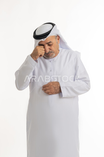 اشارات وحركات وجه ويدين مختلفة تدل على الانزعاج وعدم الرضا، بورتريه لرجل عربي خليجي إماراتي كبير بالسن يرتدي الكندورة الإماراتية التقليدية والغترة يضع قبضته على وجهه بإيماءات التألم، الوقوف والشعور بالاضطراب والتعب، خلفية بيضاء