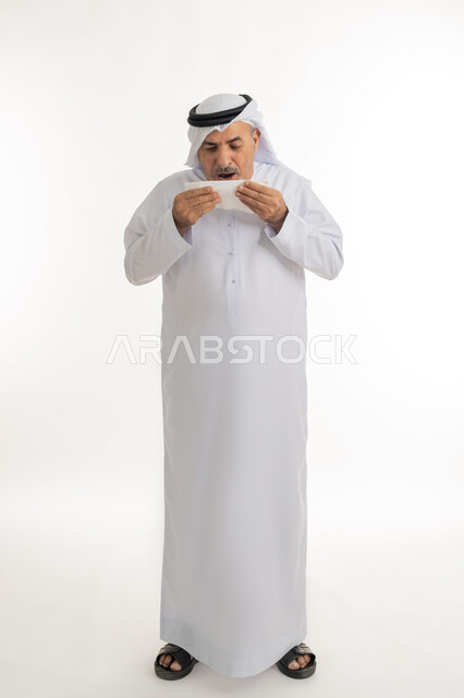 ضرورة الحصول على الراحة وأخذ العلاج المناسب, العطاس والإحساس بأعراض الزكام والإصابة بالانفلونزا، إيماءات الألم والتعب , بورتريه لرجل كبير السن عربي خليجي اماراتي يرتدي الغترة والكندورة يمسك المنديل بيديه ويغطي أنفه، الطول الكامل للجسم، خلفية بيضاء