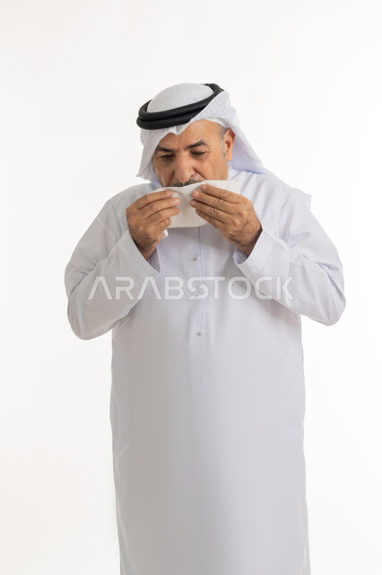 التهاب الأنف التحسسية وظهور أعراض الأنفلونزا, بورتريه لرجل كبير بالسن عربي اماراتي خليجي يرتدي الغترة والكندورة يضع يديه على وجهه بإيماءات العطاس, استخدام المناديل الورقيه للعطس, الشعور بالمرض و الزكام والحاجة الى العلاج والدواء, خلفية بيضاء