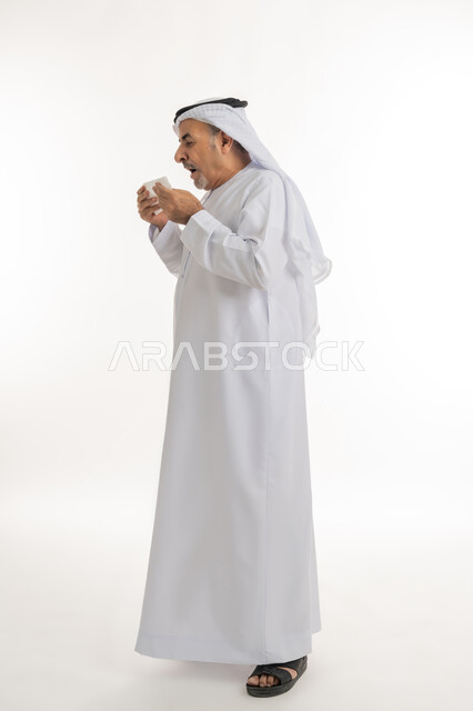 العطاس والإحساس بأعراض الزكام والإصابة بالانفلونزا، إيماءات الألم والتعب, بورتريه من الجانب لرجل كبير السن عربي خليجي اماراتي يرتدي الغترة والكندورة يمسك المنديل بيديه ويغطي أنفه، ضرورة الحصول على الراحة وأخذ العلاج المناسب, الطول الكامل للجسم، خلفية بيضاء