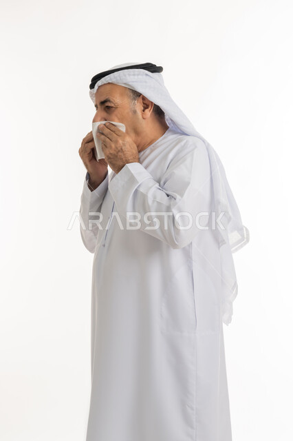 Gestures of pain, fatigue and illness, side portrait of an elderly Arab Gulf Emirati man wearing a ghutra and kandura holding a handkerchief in his hands and covering his nose, the necessity of getting rest and taking appropriate treatment, sneezing and feeling the symptoms of a cold and flu, white background