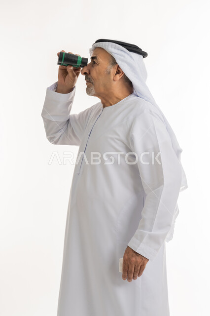 متعة الاستكشاف والمغامرة، رؤى مستقبلية عند الراشدين والكبار بالسن، بورتريه لرجل مسن عربي خليجي إماراتي  يرتدي الكندورة والغترة يقوم بالنظر من خلال المنظار ثنائي العينين، التدقيق والتركيز على شيء ما وتوسيع مجال الرؤية باستخدام المنظار، خلفية بيضاء