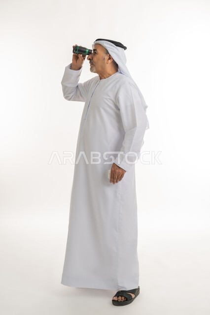 مفهوم الاستكشاف والمغامرة، رؤى مستقبلية عند الراشدين والكبار بالسن، بورتريه لرجل مسن عربي خليجي إماراتي  مبتسم يرتدي الكندورة والغترة يقوم بالنظر من خلال المنظار ثنائي العينين، البحث عن شيء ما، الطول الكامل للجسم، خلفية بيضاء
