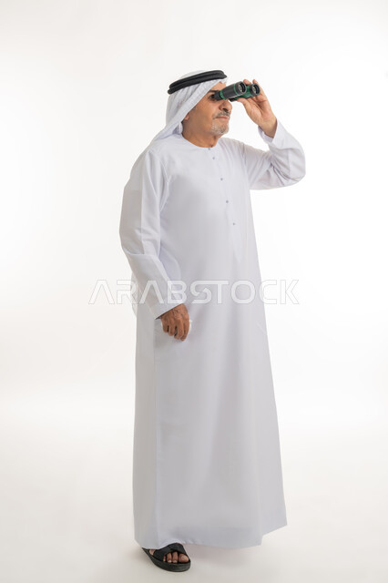رؤى مستقبلية عند الراشدين والكبار بالسن، حب الاستكشاف والمغامرة، رؤية الأشياء البعيدة، بورتريه لرجل مسن عربي خليجي إماراتي  مبتسم يرتدي الكندورة والغترة التقليدية يقوم بالنظر من خلال المنظار ثنائي العينين(التلسكوب)، صورة الجسم بالكامل، خلفية بيضاء