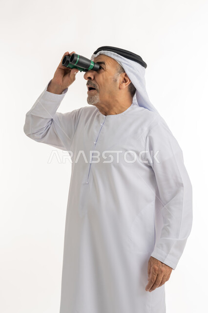 ايماءات وتعابير الدهشة والمفاجئة، رؤية الاشياء البعيدة عبر التلسكوب، رؤى مستقبلية عند الراشدين والكبار بالسن، بورتريه لرجل مسن عربي خليجي إماراتي  مبتسم يرتدي الكندورة والغترة ينظر من خلال المنظار ثنائي العينين، حب المغامرة والاستكشاف، خلفية بيضاء