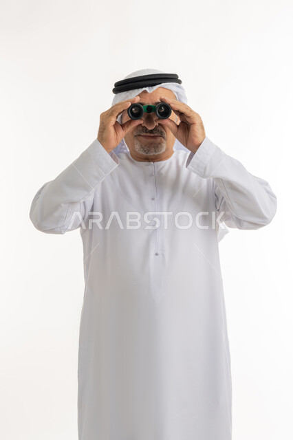 البحث عن شيء ما وتوسيع مجال الرؤية باستخدام المنظار، مفهوم الاستكشاف والمغامرة، رؤى مستقبلية عند الراشدين والكبار بالسن، بورتريه لرجل مسن عربي خليجي إماراتي  مبتسم يرتدي الكندورة والغترة التقليدية يقوم بالنظر من خلال المنظار ثنائي العينين، خلفية بيضاء