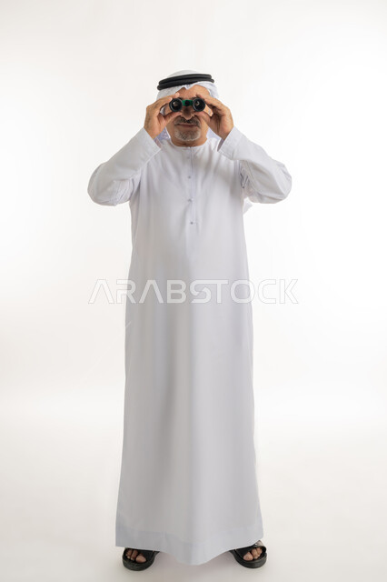 رؤية الأشياء البعيدة، حب الاستكشاف والمغامرة، بورتريه لرجل مسن عربي خليجي إماراتي  مبتسم يرتدي الكندورة والغترة التقليدية يقوم بالنظر من خلال المنظار ثنائي العينين(التلسكوب)، رؤى مستقبلية عند الراشدين والكبار بالسن، صورة الجسم بالكامل، خلفية بيضاء