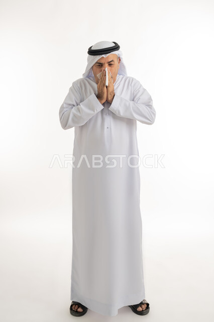 الإصابة بالانفلونزا والعطاس والإحساس بأعراض الزكام، إيماءات الألم والتعب والمرض, بورتريه  لرجل كبير السن عربي خليجي اماراتي يرتدي الغترة والكندورة يمسك المنديل بيديه ويغطي أنفه، الحاجة للراحة وأخذ العلاج المناسب, الجسم بالطول الكامل، خلفية بيضاء