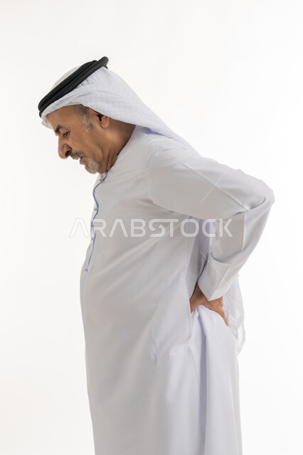 الشعور في الالم بالعمود الفقري والإصابة بمرض الديسك، آلام الظهر والعمود الفقري والتهاب المفاصل، بورتريه من الجانب لرجل عربي خليجي إماراتي كبير بالسن يرتدي الكندورة الإماراتية والغترة يقف بصعوبة ويمسك ظهره بيديه بإيماءات المعاناة، خلفية بيضاء