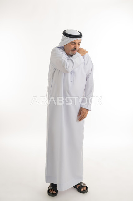إيماءات الألم في الجسد، التهاب العضلات والمفاصل، بورتريه لرجل مسن عربي اماراتي خليجي يرتدي الكندورة و الغترة يضع يده على كتفه بتعابير الوجع، الحاجه لوضع خطة علاجية, طلب المساعده للذهاب الى المستشفى، الإحساس بالتعب والإرهاق, الطول الكامل للجسم، خلفية بيضاء