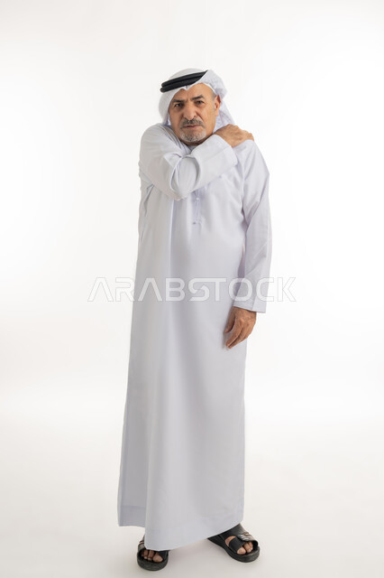طلب المساعده للذهاب الى المستشفى، الإحساس بالتعب والإرهاق, إيماءات الألم في الجسد، التهاب العضلات والمفاصل، بورتريه لرجل مسن عربي اماراتي خليجي يرتدي الكندورة و الغترة يضع يده على كتفه بتعابير الوجع، الحاجه لوضع خطة علاجية, الطول الكامل للجسم، خلفية بيضاء