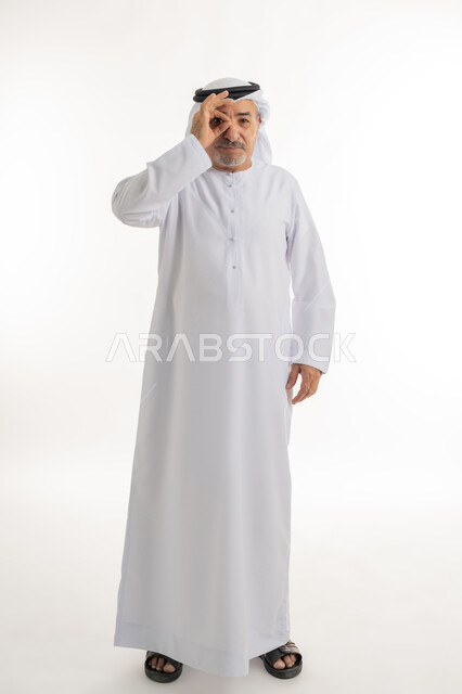 مفهوم التخطيط و الرؤية المستقبلية، بورتريه لرجل كبير بالسن عربي خليجي اماراتي يرتدي الكندورة والغترة يضع يده على عينيه بشكل عدسة بإيماءات البحث والاستكشاف، التركيز والنظر إلى شيء محدد، محاولة تقريب الأشياء البعيدة, الطول الكامل للجسم، خلفية بيضاء