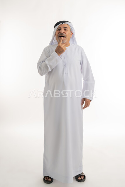 الاشارة إلى الفم بإيماءات الانزعاج والألم, مشاكل التهاب اللثة, طلب العلاج والمساعدة, الحاجة لزيارة طبيب الأسنان, بورتريه لرجل كبير بالسن عربي خليجي اماراتي يرتدي الكندورة والغترة يضع يده على فمه, صورة الجسم بالطول الكامل, خلفية بيضاء