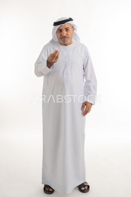الوقوف والنظر إلى الكاميرا بتعابير البشاشة، حركات وجه ويدين تدل على التروي والصبر, بورتريه لرجل كبير بالسن عربي خليجي إماراتي يرتدي الغترة والكندورة يرفع قبضته بإيماءات التمهل والانتظار، الجسم بالطول الكامل، خلفية بيضاء