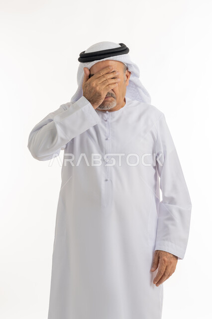 وضع اليد على الوجه وتغطيه العيون, الاندهاش والاستغراب من امر ما, تذكر شيء محدد بشكل مفاجئ, بورتريه لرجل كبير بالسن عربي خليجي اماراتي يرتدي الكندورة والغترة يقف باستقامة بإيماءات الندم, خلفية بيضاء