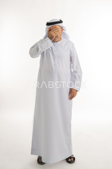 Remembering something specific suddenly, portrait of an elderly Arab Gulf Emirati man wearing a kandura and ghutra standing straight with gestures of regret, putting his hand on his face and covering his eyes, astonishment and surprise at something, full body length, white background