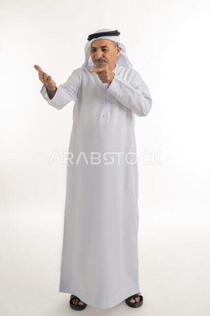 الوقوف والنظر إلى الكاميرا بإيماءات البشاشة, الإشارة إلى شيء ما في كف اليد، بورتريه لرجل كبير بالسن عربي خليجي إماراتي يرتدي الغترة والكندورة الإماراتية يحمل عنصر ما ويشير إليه بإصبع السبابة، الجسم بالطول الكامل،  خلفية بيضاء