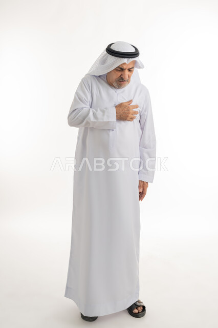 ضرورة الذهاب للطبيب والحصول على العلاج، إيماءات الوجه تدل على الاضطراب والتألم، بورتريه لرجل كبير السن عربي خليجي إماراتي يرتدي الغترة والكندورة يضع يده على صدره بإيماءات الألم, الشعور بآلام في القلب وضيق في التنفس، الجسم بالطول الكامل، خلفية بيضاء