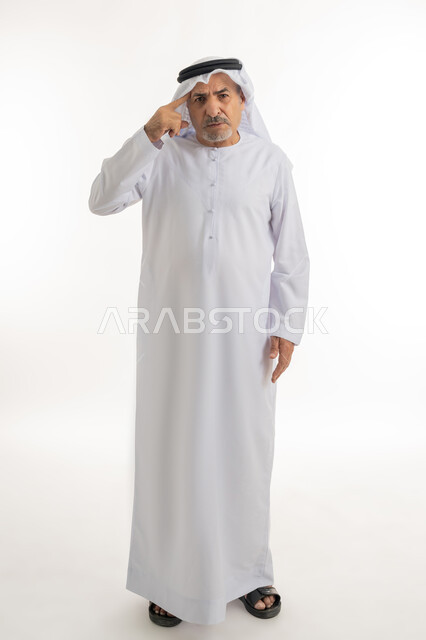 الوقوف والنظر إلى الكاميرا بإيماءت العصبية والغضب، بورتريه لرجل عربي خليجي إماراتي كبير بالسن يرتدي الكندورة والغترة الإماراتية يشير بإصبعه إلى رأسه، رجاحة العقل والتفكير،  تعابير الجدية والحزم، الطول الكامل للجسم، خلفية بيضاء