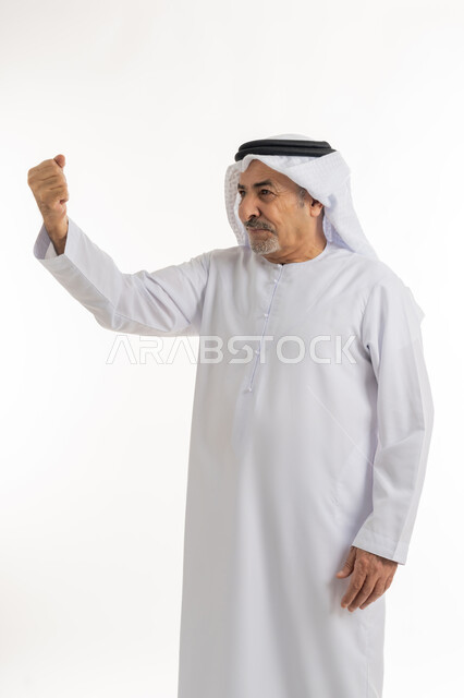 الوقوف بظهر مستقيم ومشدود والنظر الى شيء ما بإيماءات الجدية، بورتريه لرجل كبير بالسن عربي خليجي إماراتي يرتدي الكندورة والغترة يرفع يده بتعابير القوة والعزيمة، حركات وإشارات يدين تدل على التحدي والحماس، خلفية بيضاء