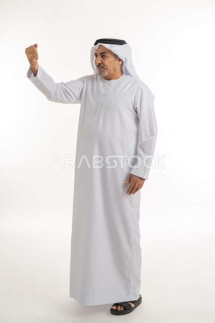 حركات وإشارات يدين تدل على التحدي والحماس، الوقوف بظهر مستقيم ومشدود والنظر الى شيء ما بإيماءات الجدية، بورتريه لرجل كبير بالسن عربي خليجي إماراتي يرتدي الكندورة والغترة يرفع يده بتعابير القوة والعزيمة، الطول الكامل للجسم، خلفية بيضاء
