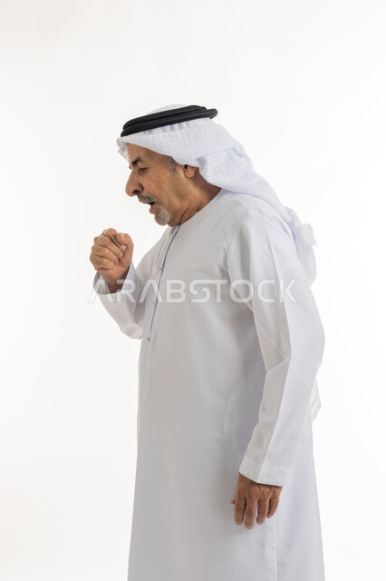 الحاجة للراحة وأخذ العلاج المناسب, الإصابة بالتهاب الحلق وأمراض البلعوم، السعال بشكل متكرر بإيماءات الألم والتعب, بورتريه من الجانب لرجل كبير السن عربي خليجي اماراتي يرتدي الغترة والكندورة يغطي فمه بيديه ويكح، خلفية بيضاء