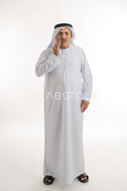 الشعور بالتعب والمرض, بورتريه لرجل كبير السن عربي خليجي إماراتي يرتدي الغترة والكندورة يضع يديه على خده بتعابير الألم, الإحساس بوجع الأسنان والتألم من النخور، الحاجة للعلاج والذهاب للعيادة السنية، الجسم بالطول الكامل، خلفية بيضاء