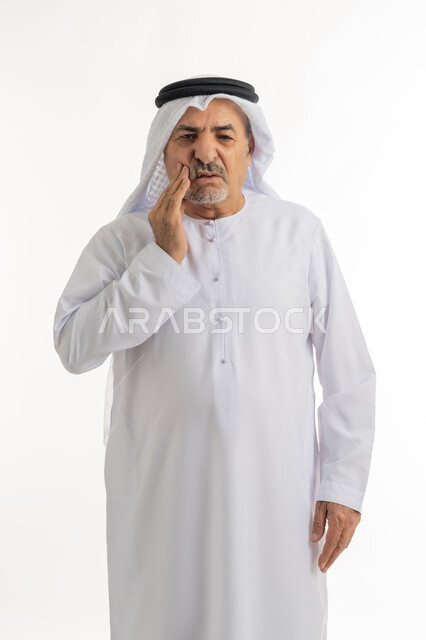 التألم من النخور ووجع الأسنان، الحاجة للعلاج والذهاب للعيادة السنية، الشعور بالتعب والمرض, بورتريه لرجل كبير السن عربي خليجي إماراتي يرتدي الغترة والكندورة يضع يديه على خده بتعابير الألم, خلفية بيضاء