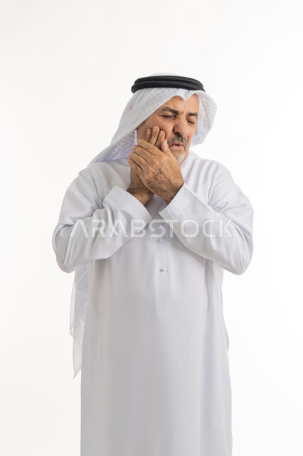 الإحساس بوجع الأسنان والتألم من النخور، الحاجة للعلاج والذهاب للعيادة السنية، الشعور بالتعب والمرض, بورتريه لرجل كبير السن عربي خليجي إماراتي يرتدي الغترة والكندورة يضع يديه على خده بتعابير الألم, خلفية بيضاء