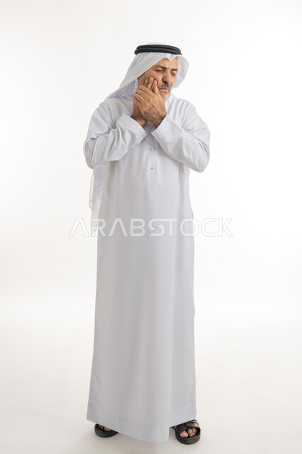 الحاجة للعلاج والذهاب للعيادة السنية، الشعور بالتعب والمرض, بورتريه لرجل كبير السن عربي خليجي إماراتي يرتدي الغترة والكندورة يضع يديه على خده بتعابير الألم, الإحساس بوجع الأسنان والتألم من النخور، الجسم بالطول الكامل، خلفية بيضاء