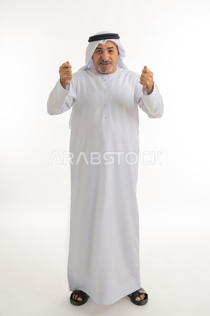 الوقوف باستقامة ورفع اليدين بإيماءات الحماس والتشجيع، إشارات وحركات تدل على العزيمة والقوة، النظر الى الكاميرا بتعابير الفرح والسعادة، بورتريه لرجل كبير بالسن عربي خليجي إماراتي يرتدي الكندورة والغترة، الطول الكامل للجسم، خلفية بيضاء