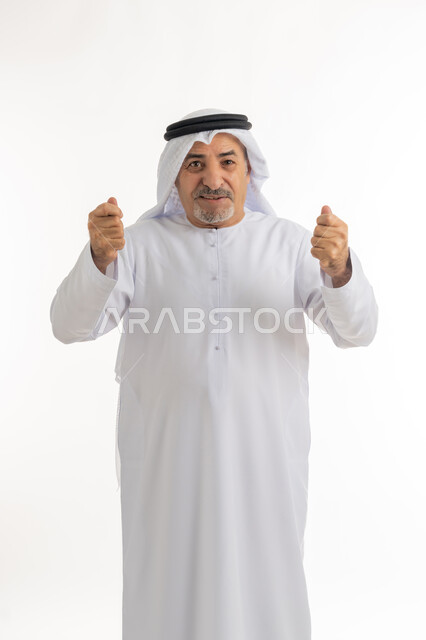إشارات وحركات تدل على العزيمة والقوة، النظر الى الكاميرا بتعابير الفرح والسعادة، بورتريه لرجل كبير بالسن عربي خليجي إماراتي يرتدي الكندورة والغترة، الوقوف باستقامة ورفع اليدين بإيماءات الحماس والتشجيع، خلفية بيضاء
