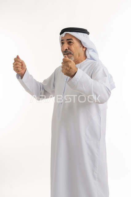 رفع اليدين بإيماءات الحماس والتشجيع، بورتريه لرجل كبير بالسن عربي خليجي إماراتي يرتدي الكندورة والغترة يقف بظهر مشدود مستقيم ينظرالى شيء ما بتعابير الفرح والسعادة، حركات وإشارات تدل على العزيمة والقوة، خلفية بيضاء