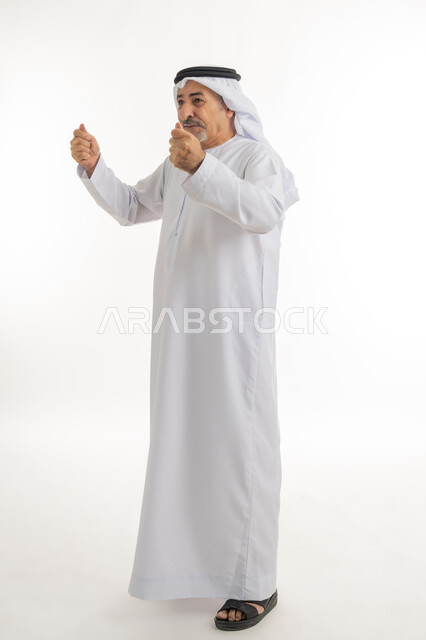 النظر الى شيءما بتعابير الفرح والسعادة، بورتريه لرجل كبير بالسن عربي خليجي إماراتي يرتدي الكندورة والغترة، الوقوف باستقامة ورفع اليدين بإيماءات الحماس والتشجيع، إشارات وحركات تدل على العزيمة والقوة، الطول الكامل للجسم، خلفية بيضاء
