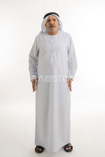 تعابير العبوس والانزعاج، نظرات التحدي والغضب، بورتريه لرجل عربي خليجي إماراتي كبير بالسن يرتدي الغترة والكندورة الإماراتية، النظر الى الكاميرا بايماءات السخط والتذمر، مفهوم عدم القبول والرضا، الشعور بالعصبية والضجر، الطول الكامل للجسم، خلفية بيضاء