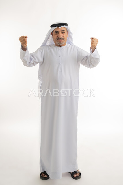 رفع اليدين بإيماءات الحماس والتشجيع، بورتريه لرجل كبير بالسن عربي خليجي إماراتي يرتدي الكندورة والغترة يقف بظهر مشدود مستقيم وينظر للكاميرا بتعابير الفرح والسعادة، حركات وإشارات تدل على العزيمة والقوة، الطول الكامل للجسم، خلفية بيضاء