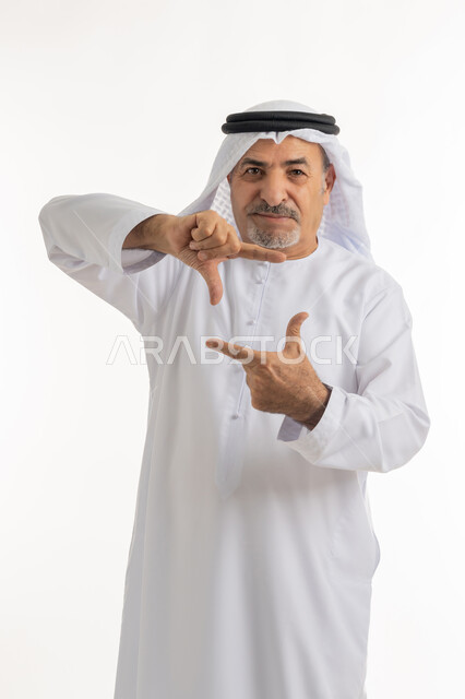 تعابير وحركات يدين تدل على التقاط صور، توثيق اللحظات السعيدة, مفهوم الرجولة والثقة بالنفس، بورتريه لرجل مسن عربي خليجي إماراتي يرتي الكندورة والغترة ينظر الى الكاميرا بايماءات البشاشة والسرور، الاهتمام بالأناقة والمظهر الخارجي، خلفية بيضاء
